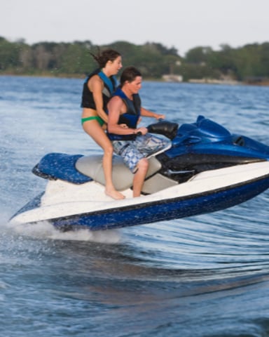 Two people riding a blue and white jet ski in shallow ocean water near a tree-lined shore