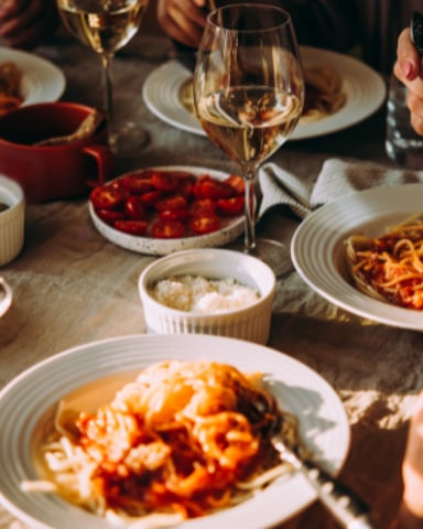 Overhead view of dinner table with pasta dishes, wine glasses, and diners' hands reaching for food