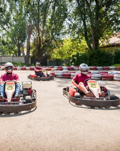 Go-kart racing track with multiple drivers in red and white karts navigating between red and white painted tire obstacles, surrounded by tall trees.