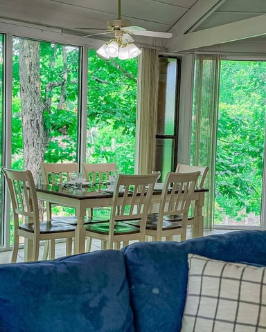 Screened porch with blue sofa, dining table with four chairs, and expansive forest views through windows
