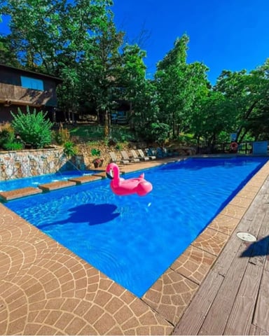 Backyard swimming pool with pink flamingo float, surrounded by lounge chairs, stone walls, and modern house with trees.