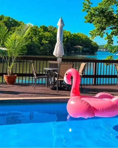 Pink flamingo pool float in swimming pool with waterfront deck, umbrellas, and lake view beyond