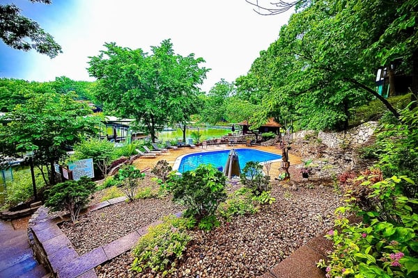 Backyard pool surrounded by lush green trees, landscaping, and stone patio with lake view beyond