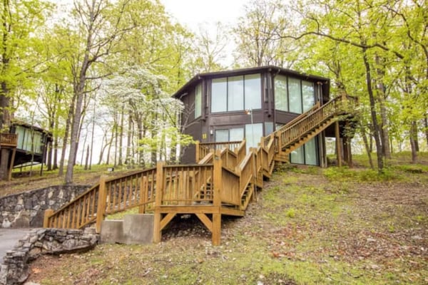 Modern elevated cabin with brown exterior and large windows surrounded by tall trees, wooden deck and stairs leading to entrance.