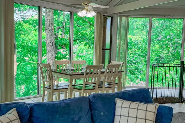 Screened porch with blue sofa, dining table, chairs, and expansive views of green forest and trees through windows
