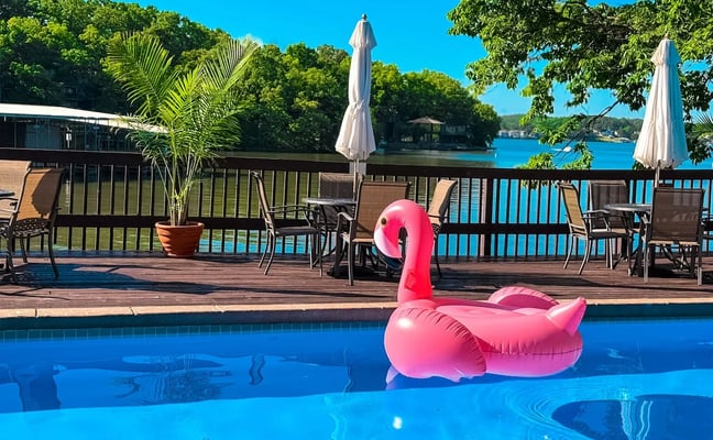 Pool with pink flamingo float, wooden deck with lounge chairs and umbrellas, lake view beyond railing