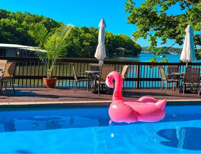 Pink flamingo pool float in swimming pool with waterfront deck, umbrellas, and lake view beyond